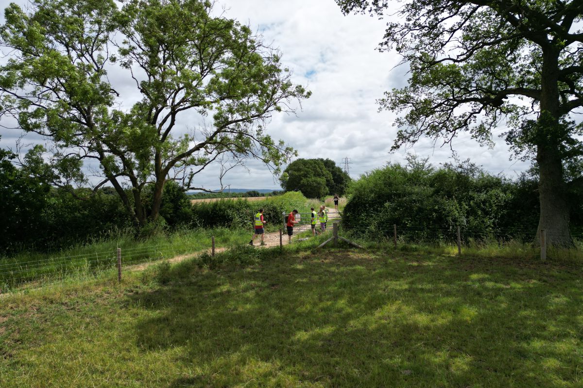 Marshals at a field gate on the course waving a runner through, with farmland beyond
